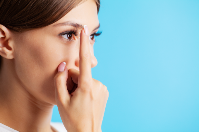 Kobieta trzyma soczewkę na palcu. Przykłada soczewkę do oka. Niebieskie tło.