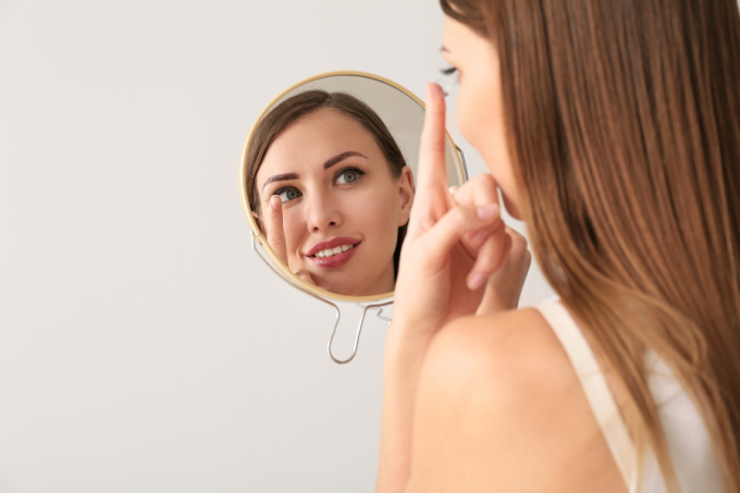 Rodzaje soczewek miękkie vs soczewki twarde, młoda kobieta zakłada soczewkę na oko przed lustrem